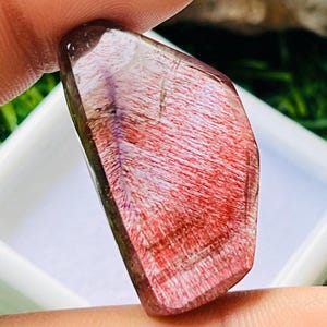 Puede incluir: Una piedra preciosa pulida con un tono marrón rojizo e inclusiones internas en forma de cabello. La piedra se sostiene entre los dedos, mostrando su calidad translúcida y sus patrones únicos. La gema se encuentra sobre un fondo blanco.
