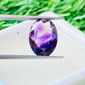 Peut inclure: Une pierre précieuse de forme ovale, aux teintes violettes, bleues et claires, maintenue par une pince argentée. La gemme est présentée dans un récipient blanc carré, sur fond d'herbe verte.