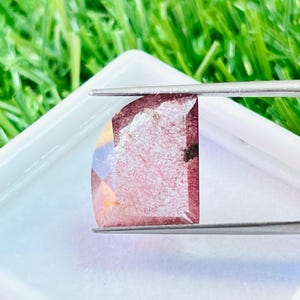 Peut inclure: Une pierre précieuse facettée, tenue par une pince, présentant un mélange de teintes roses, rouges et claires. La gemme est posée sur un fond blanc dans un récipient carré, avec de l'herbe verte en arrière-plan.