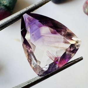 May include: A triangular-shaped, faceted gemstone held by tweezers. The gem features a gradient of colours, including purple, clear, and red. The gem is held by silver tweezers, with other stones in the background.