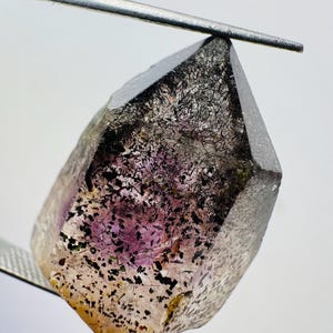 Peut inclure: Un cristal facetté à pointe, maintenu par un outil métallique. Le cristal présente un dégradé de couleurs, dont le transparent, le violet et le jaune, avec des inclusions noires. Il s'agit d'une pierre précieuse naturelle.