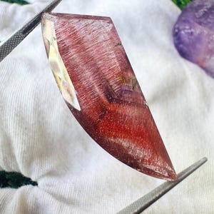 May include: A faceted, elongated gemstone with a translucent appearance, featuring deep red and maroon internal needle-like inclusions. The stone is held by metal tweezers, displaying a sharp, pointed tip and a polished, multi-faceted surface.