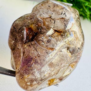 以下が含まれることがあります： 半透明の茶灰色をした、不規則な形の研磨された宝石。石の内部には、針のような黒い筋、散らばった黄金色の斑点、そして深い赤紫色の点など、目に見える内包物が含まれています。