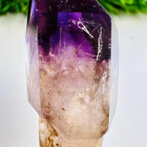 Pode incluir: Uma ponta de cristal de ametista polida com um topo translúcido de cor roxa profunda que transita para uma base mais clara e semitransparente. O cristal exibe inclusões internas naturais, incluindo pequenas manchas escuras e fissuras no corpo.