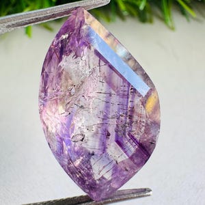 May include: A faceted, pear-shaped amethyst gemstone featuring distinct purple colour zoning and internal dark needle-like inclusions. The translucent stone displays a polished surface with sharp edges, held by metal tweezers at the top and bottom.