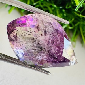 May include: A faceted pear-shaped gemstone featuring a translucent purple hue with visible dark needle-like inclusions throughout the stone. The gem is held by a pair of silver-toned metal tweezers, showcasing its internal clarity and geometric cut.
