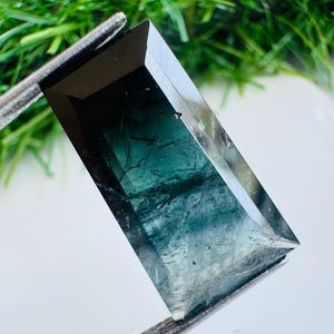 May include: A rectangular faceted gemstone with a gradient color transition from deep black at the top to a translucent teal green at the base, featuring internal inclusions and sharp beveled edges held by metal tweezers.