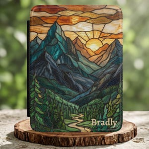 May include: A rectangular e-reader case featuring a stained glass style print of a mountain landscape at sunset. The design includes dark blue and green mountains, a winding path through a forest, and a yellow sun. The name Bradley is printed in white.
