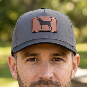 May include: A charcoal gray baseball cap featuring a rectangular brown leather patch on the front. The patch displays a black silhouette of a dog standing in profile. The cap has a structured crown, a curved brim, and a button on the top center.