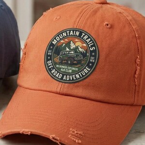 May include: Two distressed baseball caps, one blue and one orange, each featuring a circular patch on the front. The patch displays a graphic of a vehicle in a mountain landscape with the text Mountain Trails Off-Road Adventure Wilderness Explorers.