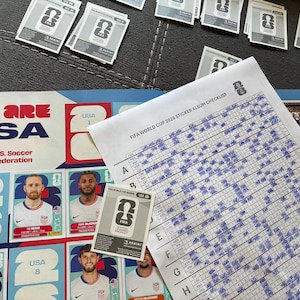 May include: A FIFA World Cup 2026 sticker album page featuring U.S. Soccer Federation branding, player portraits, and a checklist sheet with blue ink markings. Several loose Panini stickers are scattered across the top and center area.