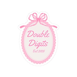 May include: A pink sticker with the text "Double Digits" in a script font, inside a pink oval frame with a scalloped edge. A pink bow is at the top. The text "Est 2026" is at the bottom of the oval.
