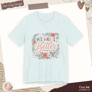 Puede incluir: Una camiseta de manga corta de color azul claro con un gráfico central que incluye el texto "His Way is Better" y "Proverbs 3:5-6". El diseño está rodeado por una corona floral con flores naranjas y blancas, hojas verdes y estrellas de mar.