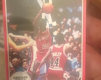 May include: A vintage basketball trading card featuring Michael Jordan of the Chicago Bulls. The card has a red border and displays Jordan in action, wearing his number 23 jersey. The card also includes the Chicago Bulls logo and the text "STAR '85".