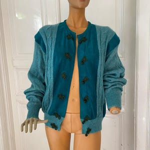 Könnte beinhalten: Türkisfarbene Strickjacke mit einzigartigem Design. Der Pullover hat eine Mittelbahn und Schulterakzente in dunklerem Türkis, verziert mit dunkelgrünen Zierverschlüssen. Die Ärmel und Seitenteile sind in einem helleren Türkis gehalten, was einen auffälligen Kontrast erzeugt.