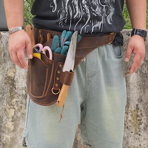Op de afbeelding: Een bruine leren tuingereedschapsriem met meerdere zakken en lussen. De riem bevat diverse gereedschappen, waaronder een troffel, een schaar en handschoenen. De riem wordt om de taille gedragen voor gemakkelijke toegang tot tuingereedschap.