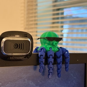 May include: A green and blue 3D-printed octopus with sunglasses perched on a laptop. The octopus has articulated blue tentacles and is positioned near a black Microsoft webcam. The webcam has the word "HD" printed on it.
