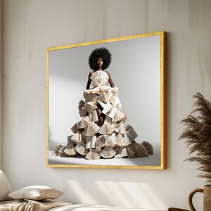 May include: A framed artwork featuring a person wearing a dress constructed from open books. The dress is beige and white, with the books fanned out to create the shape. The frame is gold, and the background is a neutral gray.