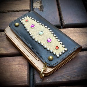 May include: A rectangular leather wallet featuring a dark brown exterior and a lighter tan base layer. The top flap is adorned with a central textured beige panel, silver-toned metal studs, five colourful circular gemstones, and a brass-toned hook.