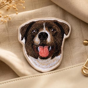 May include: An embroidered patch featuring the head of a brown and white dog with a black nose and a pink tongue sticking out. The patch has a cream-colored border and detailed stitching that captures the texture of the dog's fur and facial features.