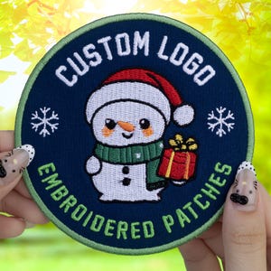 May include: Embroidered patch with a snowman design. The patch is round with a navy blue background and a green border. The snowman wears a red hat and green scarf, holding a red gift. Text reads "CUSTOM LOGO EMBROIDERED PATCHES".