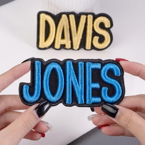 May include: Embroidered patches with the names "DAVIS" in gold and "JONES" in blue. Both patches have a black border and are rectangular in shape. The "JONES" patch is held by a person.