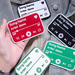 May include: A collection of rectangular embroidered patches featuring a music player interface design. Each patch displays the text Song Name and Artist Name, a progress bar, a heart icon, and playback controls in white thread on red, black, green, or white backgrounds.