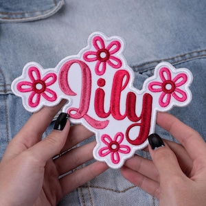 May include: Embroidered patch in the shape of a flower with the name "Lily" in pink script. The patch has a white background and is surrounded by pink flower accents. The patch is held against a denim jacket.