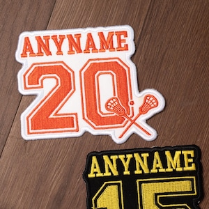 May include: Embroidered patch with the text "ANYNAME" above the number "20" in orange with a white border. Two crossed lacrosse sticks are on the right. A second patch with the text "ANYNAME" and the number "45" is below.