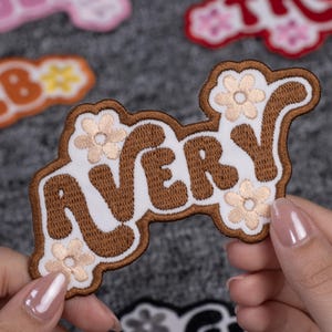 May include: Embroidered patch with the name "AVERY" in brown, outlined in a darker brown, with a white background. The patch is decorated with small, light-colored flower designs. Other patches are visible in the background.