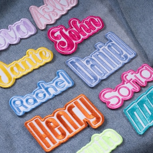 May include: A collection of embroidered name patches in various colors including pink, red, yellow, blue, orange, and teal, each featuring a white border and cursive lettering. The names displayed are John, Daniel, Rachel, Sofia, and Henry on denim.