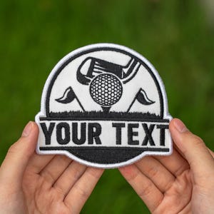 May include: A white and black embroidered patch with a golf theme. The design includes a golf ball on a tee, golf club, and flags. The text "YOUR TEXT" is displayed in bold, black letters.