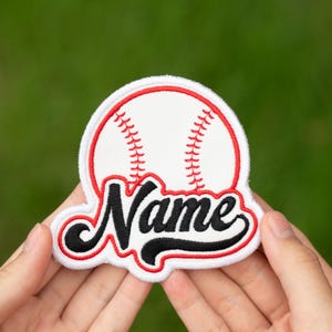 May include: A white baseball patch with red stitching and a red outline. The word "Name" is embroidered in black script across the bottom. The patch is held in front of a blurred green background.