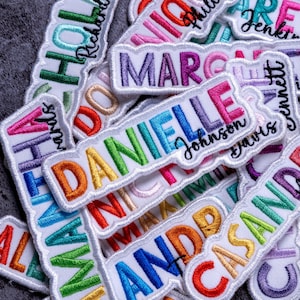 May include: A collection of embroidered name patches featuring colorful block letters and cursive script names. Each patch displays a first name in bold, multi-colored thread, accompanied by a smaller, black cursive surname stitched underneath.