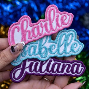 May include: Three embroidered name patches in pink, teal, and purple. The names "Charlie", "Isabelle", and "Juliana" are stitched in a cursive font with white backing. The patches are held against a backdrop of blue and green tinsel.