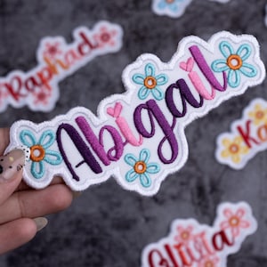 May include: Embroidered patch with the name "Abigail" in purple and pink script, surrounded by white stitching and blue and orange flower accents. Other patches with names are visible in the background.