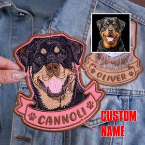 May include: A custom embroidered Rottweiler dog patch featuring a black and tan canine portrait with a pink banner underneath displaying the name CANNOLI in brown letters, accented by two small paw prints on the sides of the banner.