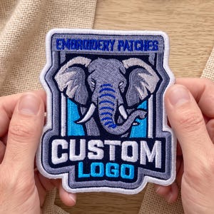 May include: A rectangular embroidered patch featuring a gray elephant head with blue accents, framed by a dark blue and white border. The text EMBROIDERY PATCHES is stitched at the top, and the words CUSTOM LOGO are stitched in white and blue at the bottom.