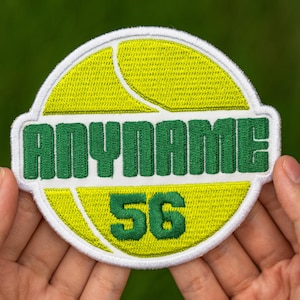 May include: Embroidered patch with a tennis ball design in yellow and white, featuring the text "ANYNAME" in green and the number "56". The patch is held in a person's hands against a blurred green background.