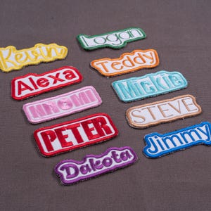 May include: A collection of ten rectangular embroidered name patches featuring the names Kevin, Logan, Alexa, Teddy, Mickie, Naomi, Steve, Peter, Jimmy, and Dakota in various thread colors including yellow, green, orange, red, pink, blue, and purple.