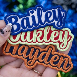 May include: Three embroidered name patches in blue, red, and orange. The blue patch says "Bailey" in white script, the red patch says "Oakley" in green script, and the orange patch says "Hayden" in dark blue script. Each patch has a contrasting border.
