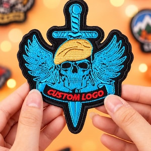 May include: An embroidered patch featuring a bright blue skull wearing a tan beret, positioned in front of a vertical sword. The design includes blue wings extending from the skull and a black banner at the bottom with the text CUSTOM LOGO in red.