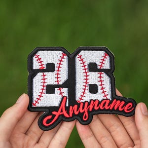 May include: A baseball-themed patch with the number "26" in a black, outlined font, with white stitching and red baseball-style stitching. The word "Anyname" is in red script below the number, also outlined in black. The patch is held in front of a green background.