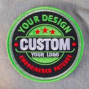 May include: A round, embroidered patch with a black background and green border. The patch features the words "YOUR DESIGN" and "CUSTOM YOUR LOGO EMBROIDERED PATCHES" in white and red lettering. Three red stars are above the word "CUSTOM".
