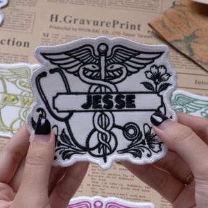 May include: A white embroidered patch featuring a black caduceus symbol, a stethoscope, and floral accents. A horizontal banner across the center contains the name JESSE in bold black letters. The patch has a scalloped edge and a white border.