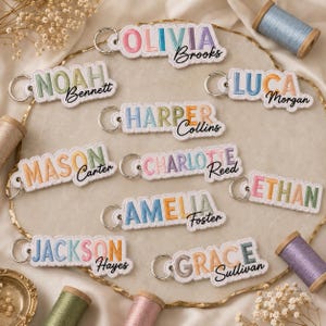 May include: A collection of rectangular embroidered keychains featuring names in colorful block letters with surnames in black cursive script. Each keychain includes a silver metal split ring. The names include Olivia Brooks, Noah Bennett, Luca Morgan, Harper Collins, Mason Carter, Charlotte Reed, Amelia Foster, Ethan, Jackson Hayes, and Grace Sullivan.