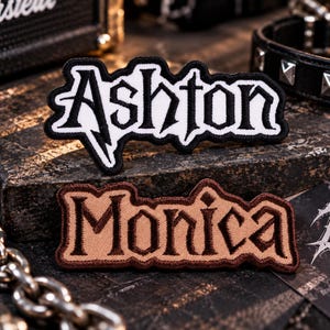 May include: Two embroidered fabric patches featuring custom names in a stylized gothic font. The top patch displays the name Ashton in white thread with a black border. The bottom patch displays the name Monica in brown thread with a dark brown border.
