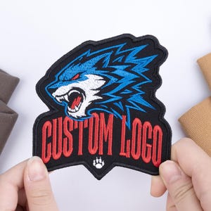 May include: An embroidered patch featuring a stylized blue and white wolf head with red eyes and an open mouth. Below the wolf, the text CUSTOM LOGO is stitched in red capital letters. A small white paw print icon is centered at the bottom edge.