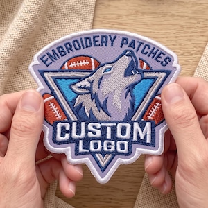 May include: A custom embroidered patch featuring a howling wolf head centered within a blue triangle. The design includes orange footballs, a light purple background, and the text EMBROIDERY PATCHES at the top and CUSTOM LOGO at the bottom.