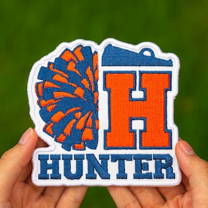May include: Embroidered patch featuring a blue and orange pom-pom design and the letter "H" in orange. The word "HUNTER" is embroidered in blue below the letter. The patch has a white border.